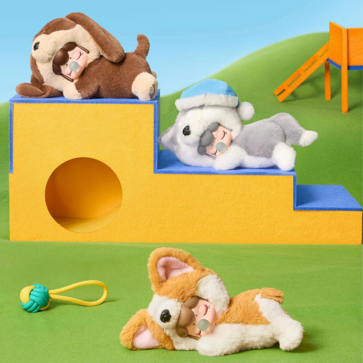 Nanci Puppy Club Dachshund, Husky and Corgi plush dolls resting on yellow-and-blue stepped playground blocks, with a rope toy on green felt grass.