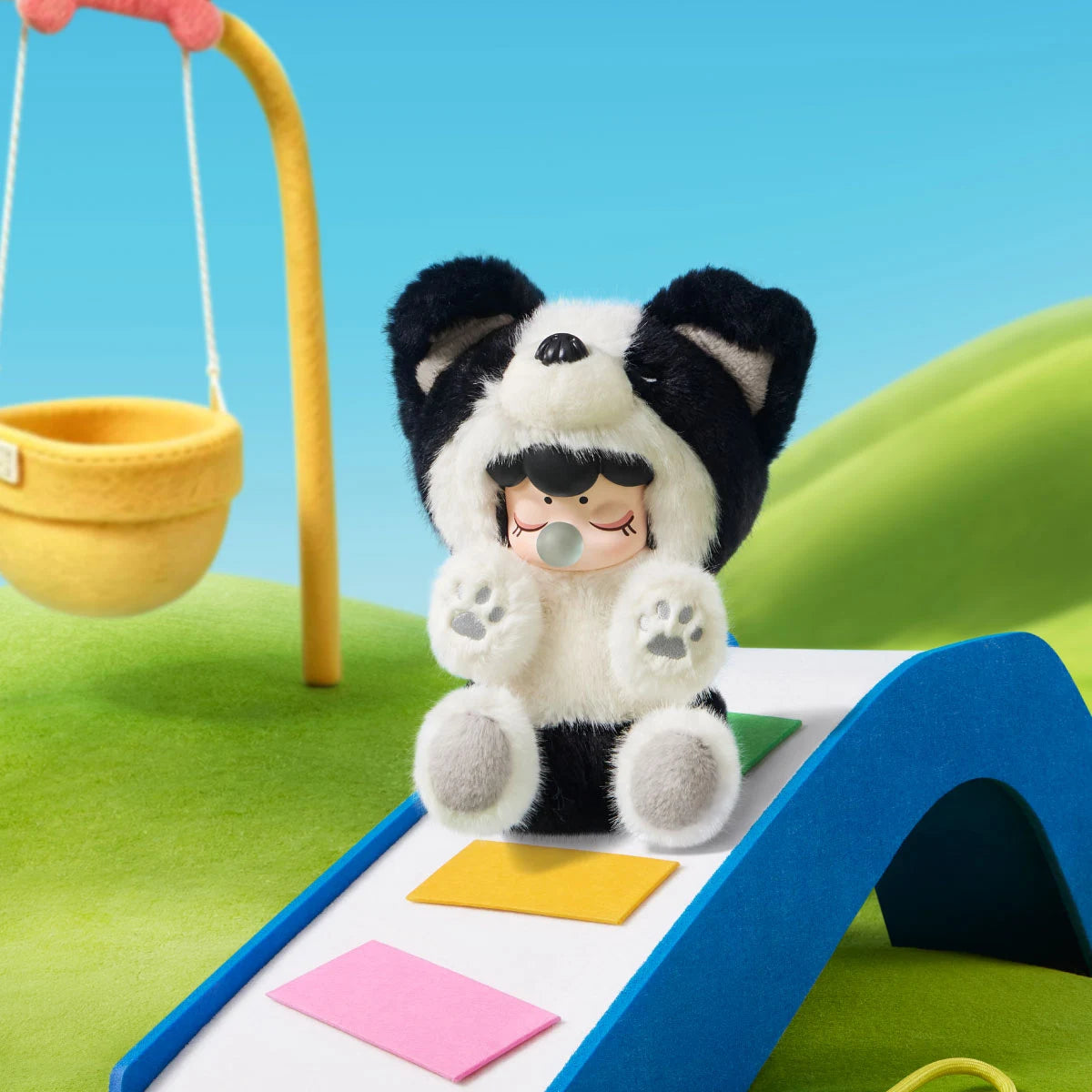 Black-and-white Nanci Puppy Club Border Collie plush doll sitting on a blue miniature slide with colorful steps, showing raised paws, a sleeping face and blue bubble.