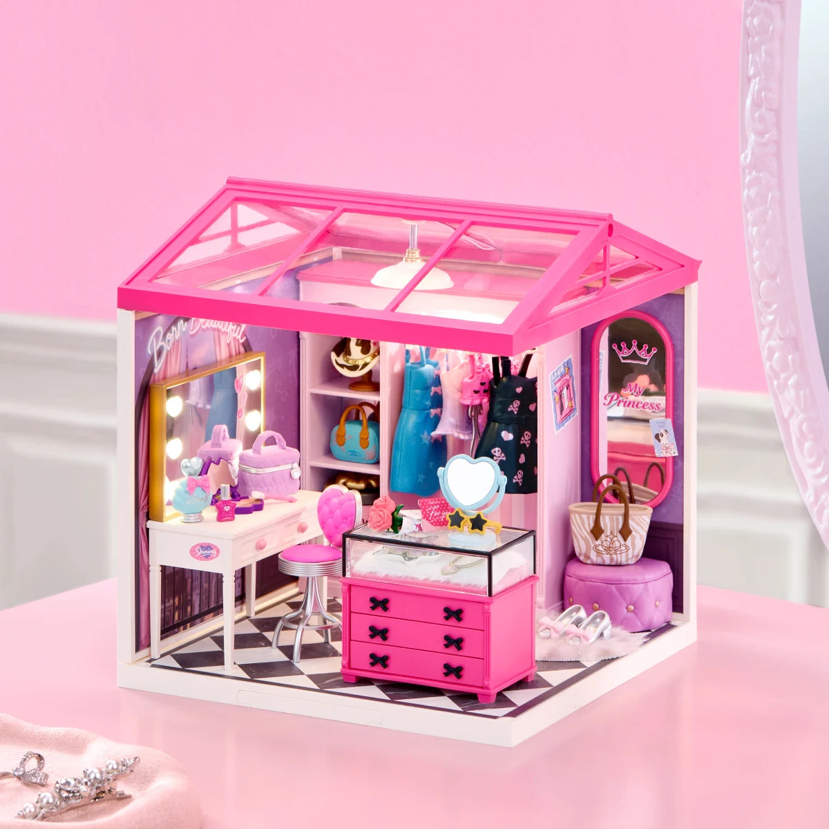 Miniature dressing room with pink glass roof, vanity, clothing rack and pink drawers displayed on a pink surface.
