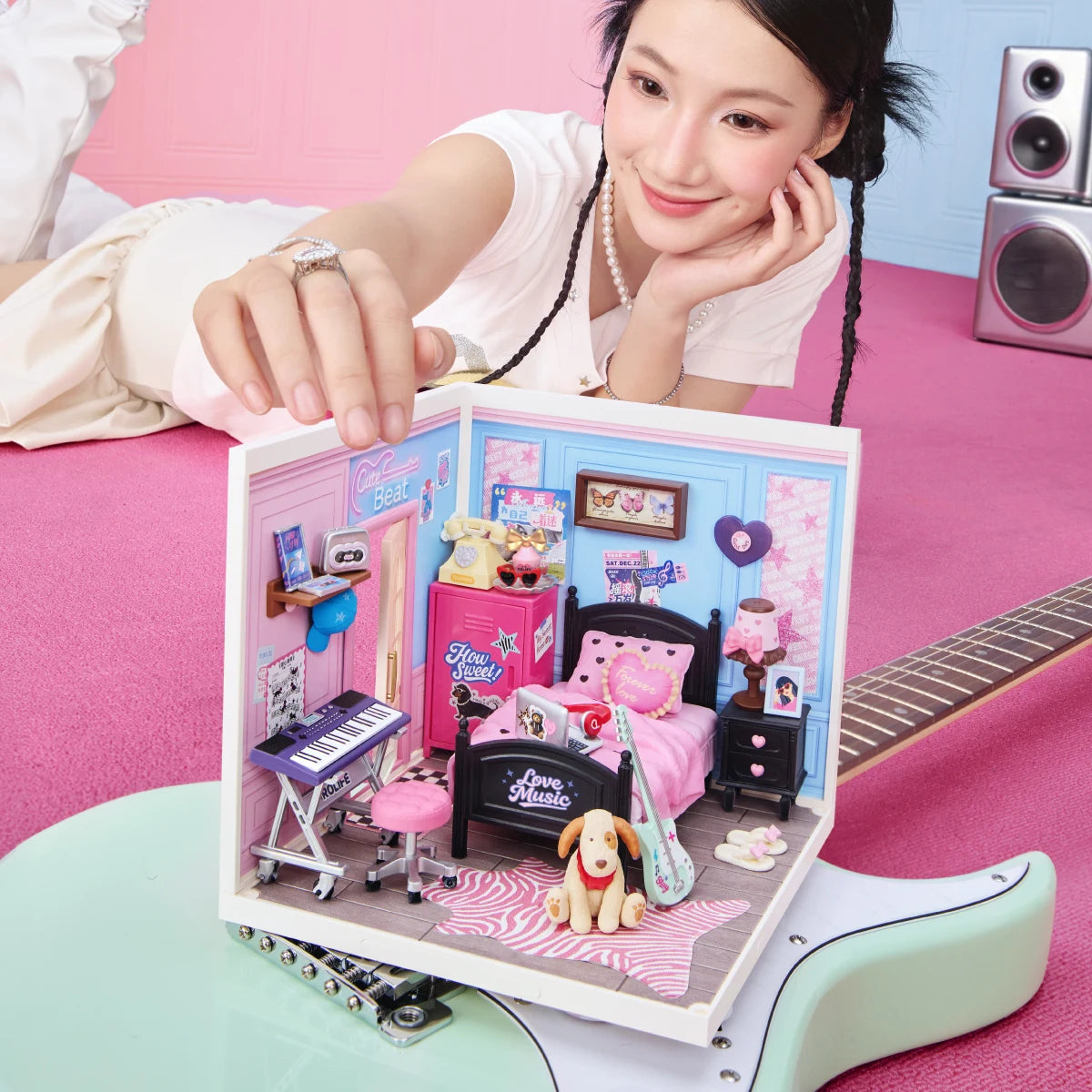 Rolife miniature bedroom diorama displayed on a pink surface, with a person's hand near the model and a guitar neck visible in the foreground.