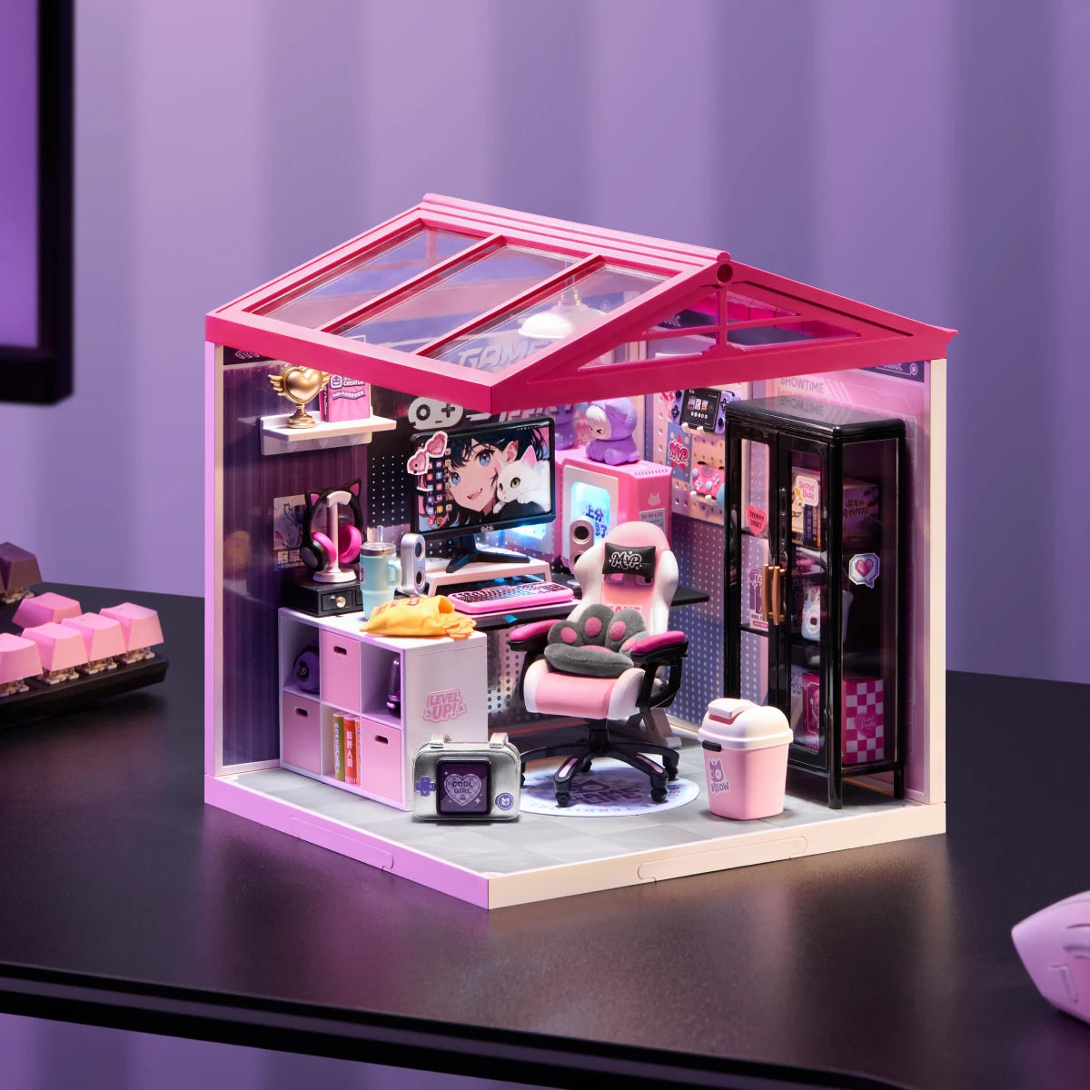 Miniature gaming room diorama with pink roof and clear panels displayed on a dark desk, showing gaming chair, desk, monitor, and cabinet.