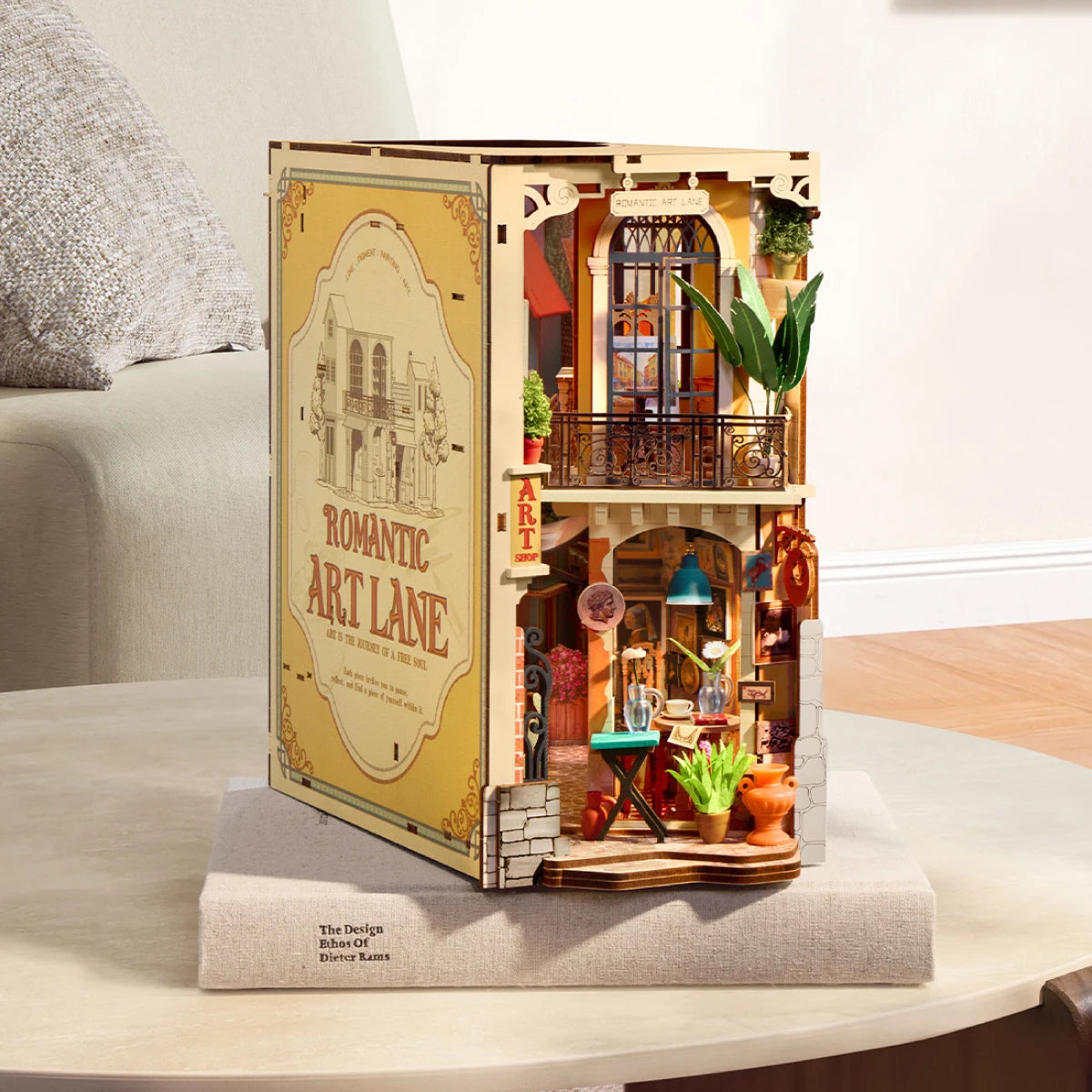 Rolife Romantic Art Lane miniature book nook standing on a light fabric-covered table beside a sofa, showing its illustrated side panel and open shop front.