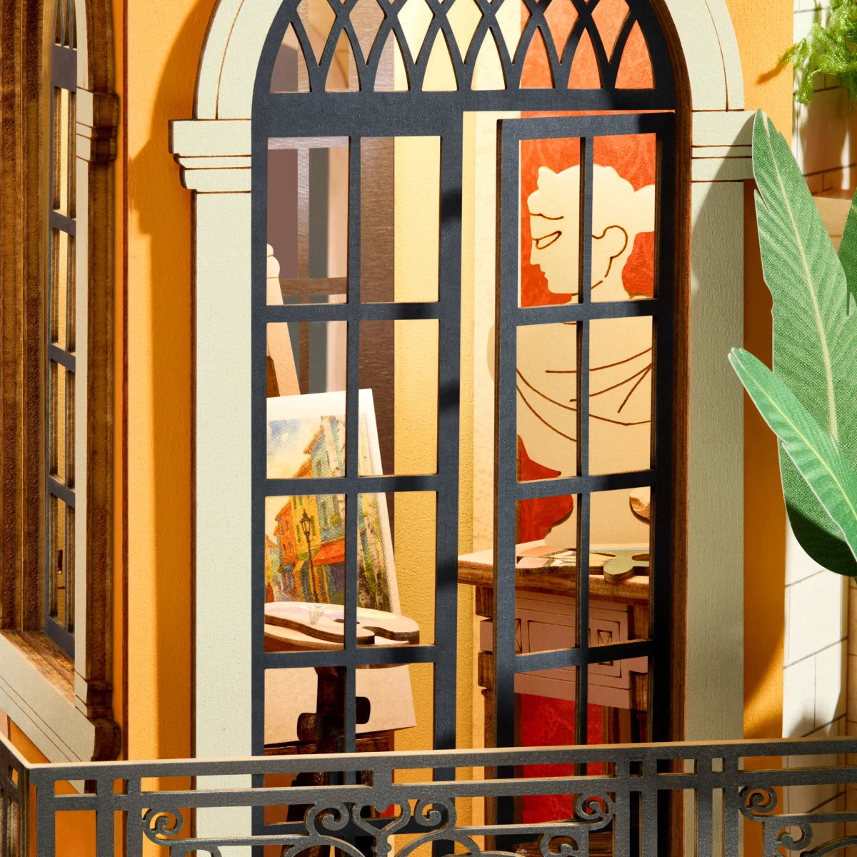 Miniature art studio seen through an arched black-framed window, with a small easel painting, line-art portrait, wooden desk and green plant leaves.