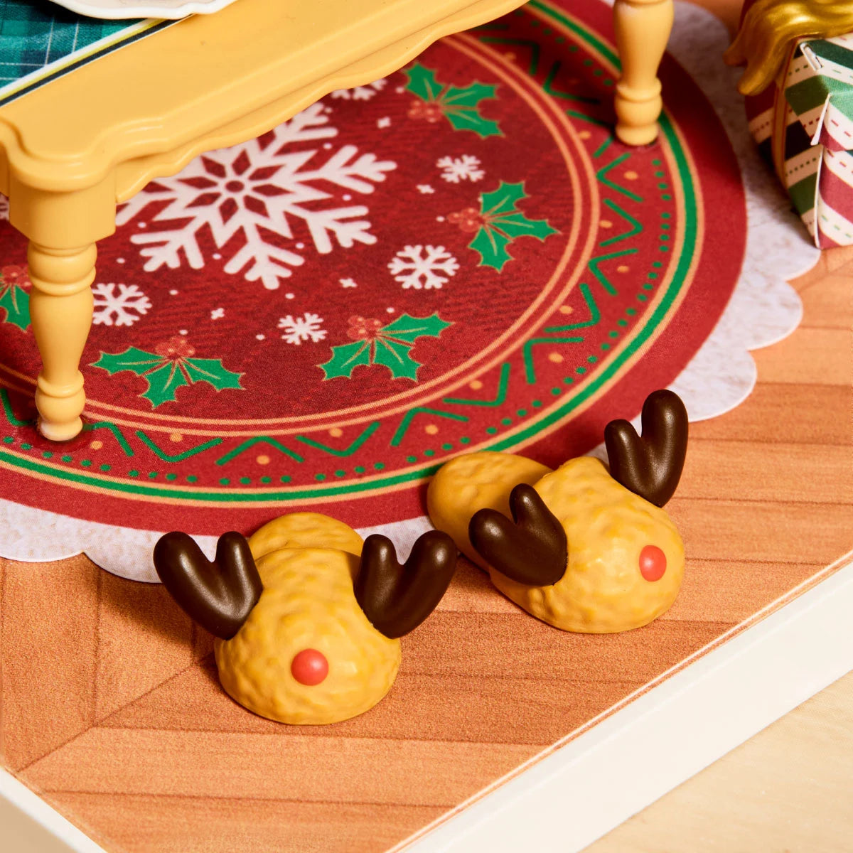 Close-up of two miniature reindeer slippers with brown antlers and red noses on a snowflake-patterned rug.